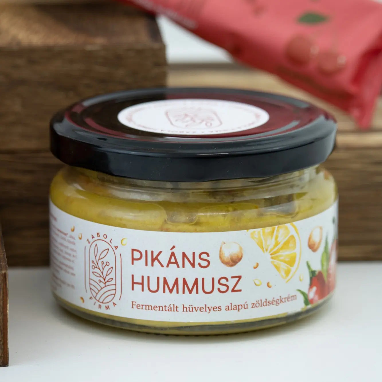 Pikán hummusz feliratú üvegcse, a kupak fekete, a hummusz nagyon szép aranysárga. A háttérben egy cseresznyeszínű csomagolás van elmosódva. 