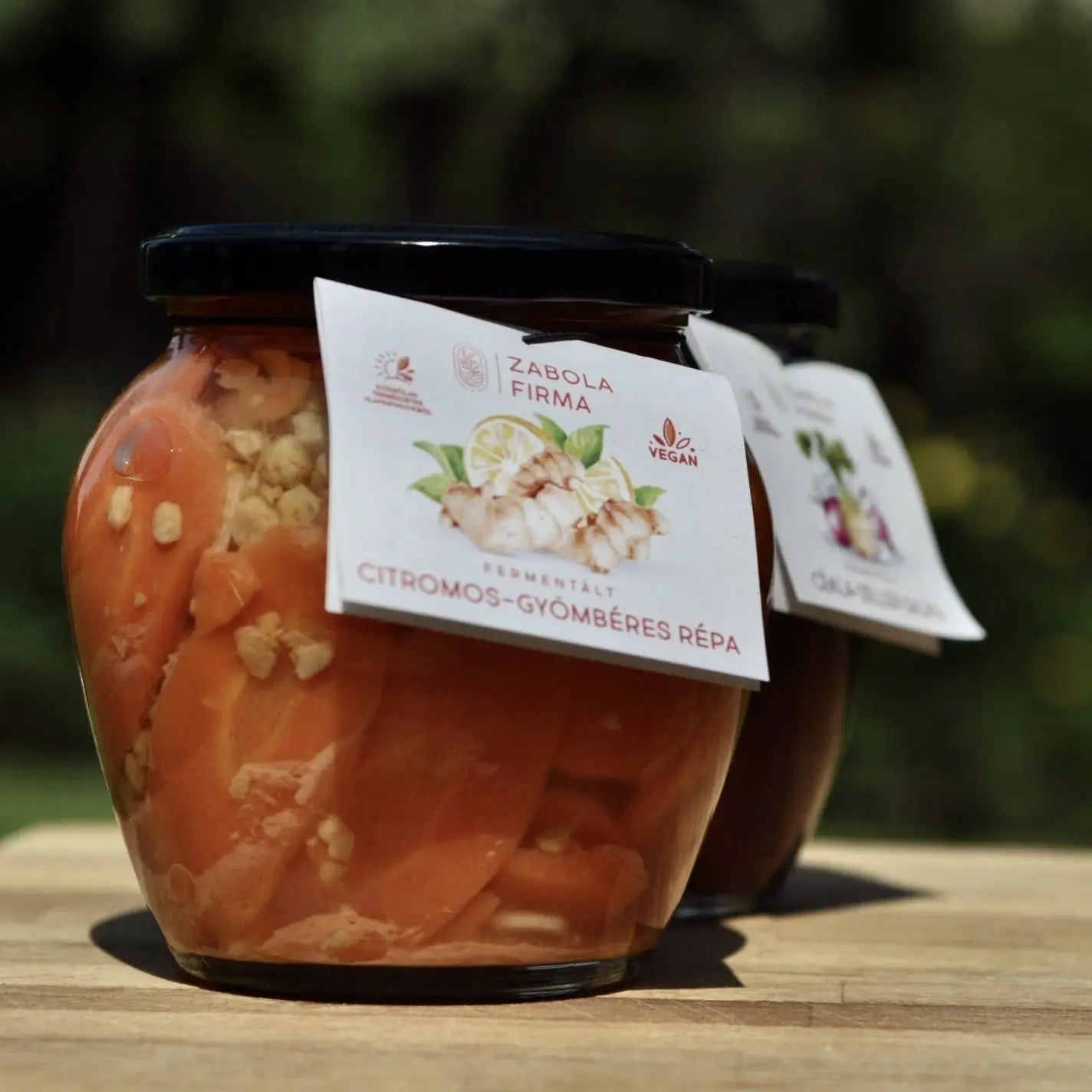 A napsütötte üvegben citromos- gyömbéres répa van, látszanak a gyömbér darabkák és a szépen felvágott répaszeltek. Az üveg egy faasztallapon áll. 