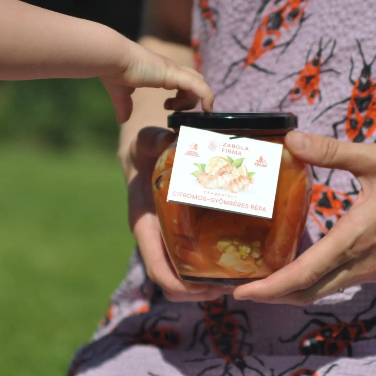 Egy lány, bodobácsos ruhában egy üveget tarta, amelyben piros színű citromos-gyömbéres répa van. Az üveg kupakja. fekete. 