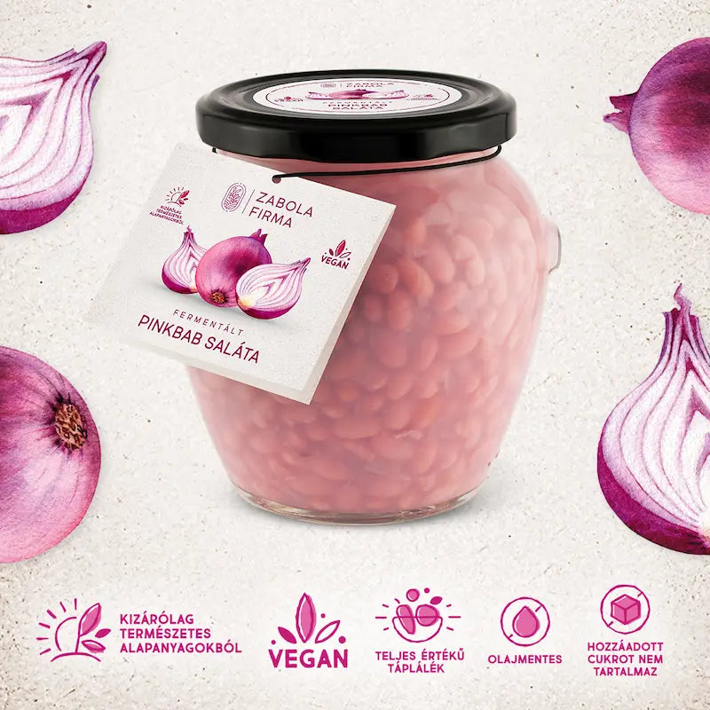 Fermentált pinkbabsaláta látható az üvegben. A babszemek nagyon gusztusak, halvány rózsaszínűek. Az üvegnek fekete kupakja van. Az üveg körül félbevágott lilahagymák vannak. 