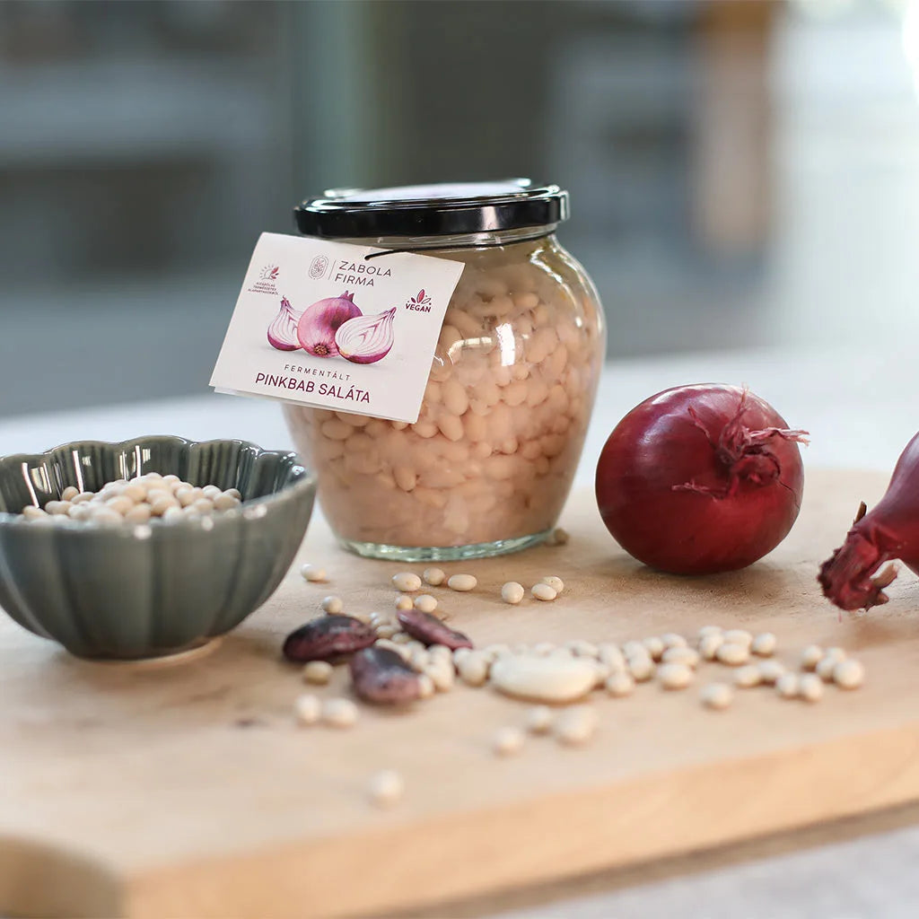 Fermentált pinkbab saláta a Lardershelf kínálatából látható a képen. A rózsaszín babok a fermentált lébenúszkálnak. Az üveg körül lilahagyma fej, babszemek találhatóak egy favágódeszkán. Az üveg mellett van egy szürke porcelántál fehér babbal. 