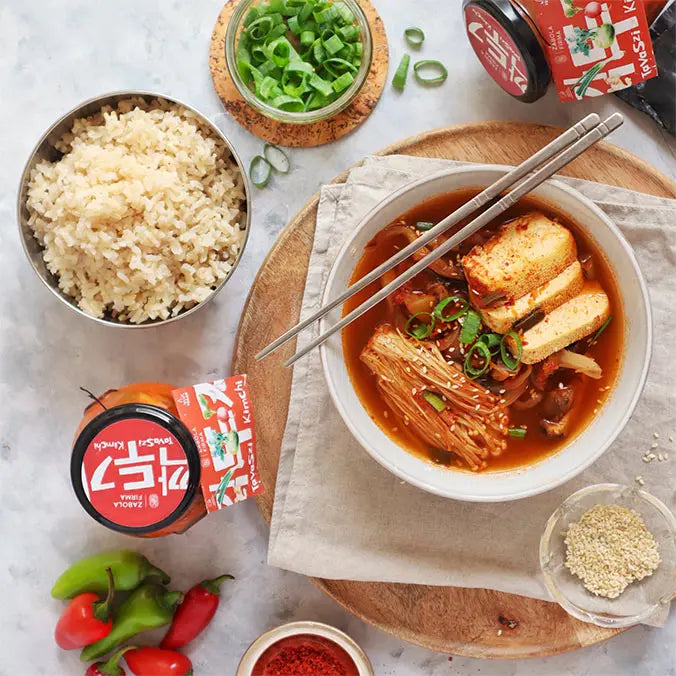 A képen egy Zabola fermentált tavaszi kimchivel készített tofus, zöldséges és tésztás ázsiai levest lehet látni. A fehér tányér mellett rizs és újhagyma karikák vannak. A tányéron két evőpálcika található. 