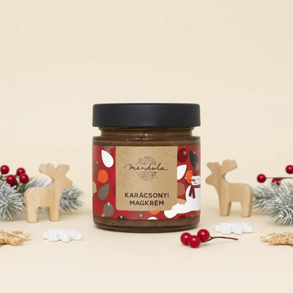 Mendula "Karácsonyi magkrém" látható a képen. Körlülette fából készült rénszarvas figurák, piros bogyók és fenyőágak. Az üvegen színes, piros, narancssárga és fehér körök, találhatóak. A kupak színe fekete. 