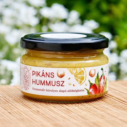 Pikáns hummusz a Lardershelf kínálatából. Az üveg lapos, fekete kupakkal van lezárva. A hummusz arany színű, a fehér címékén csicseriborsó, citrom és paprika grafikája látható. 